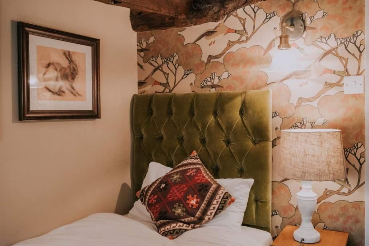 A bedroom with a tufted green headboard, a patterned pillow, a framed artwork, and a lamp on a bedside table.