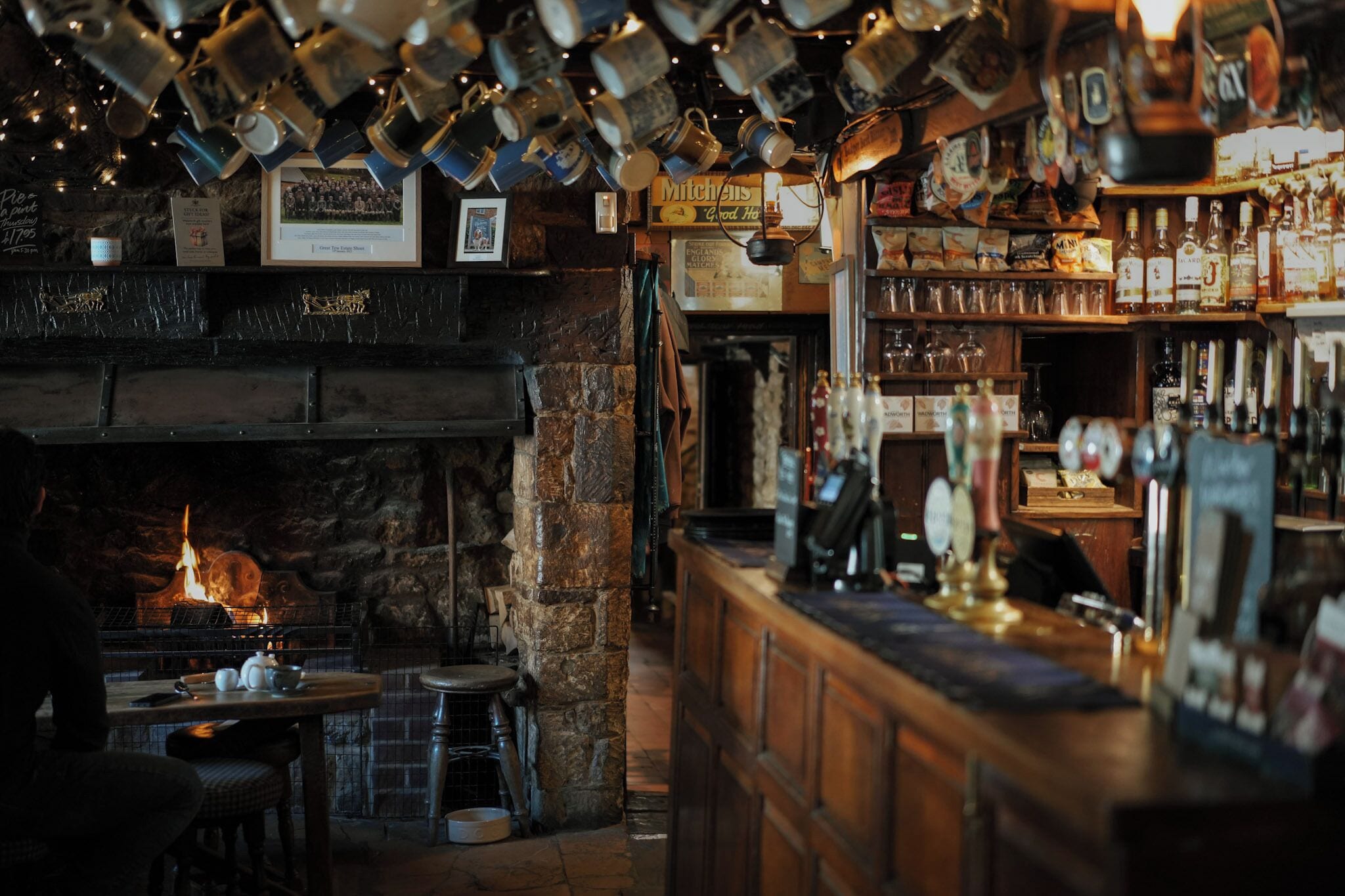 A bar area with a stone fireplace, wooden bar counter, and hanging mugs.