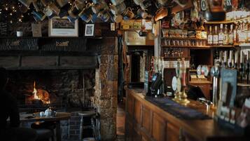 A bar area with a stone fireplace, wooden bar counter, and hanging mugs.
