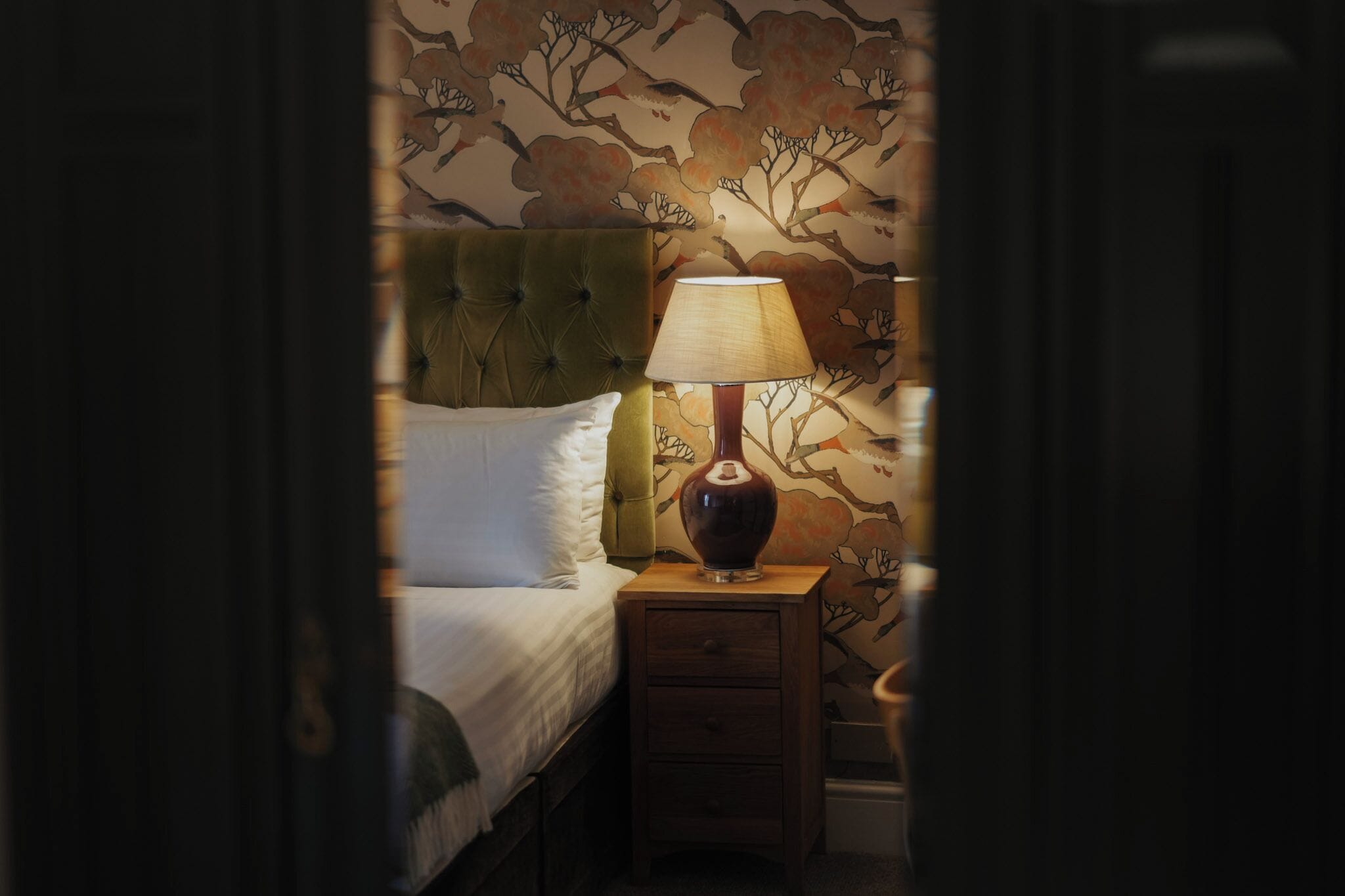 A dimly lit room with a bed, a nightstand with a lamp, and a floral wallpaper.