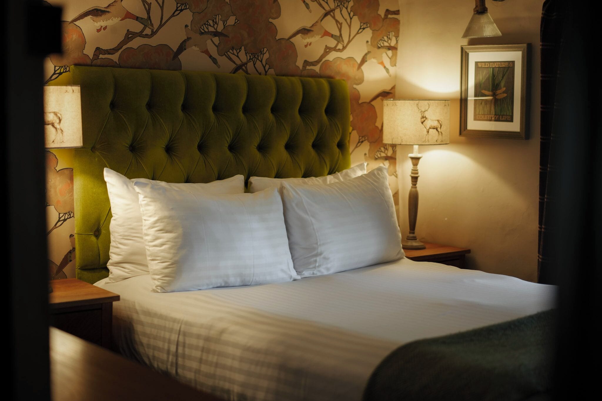 A neatly made bed with white linens and a green upholstered headboard. There are two bedside lamps and a framed picture on the wall.