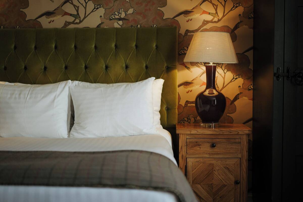 A bed with a tufted headboard, white pillows, and a plaid blanket. There is a wooden nightstand with a lamp and a wooden cabinet with drawers.