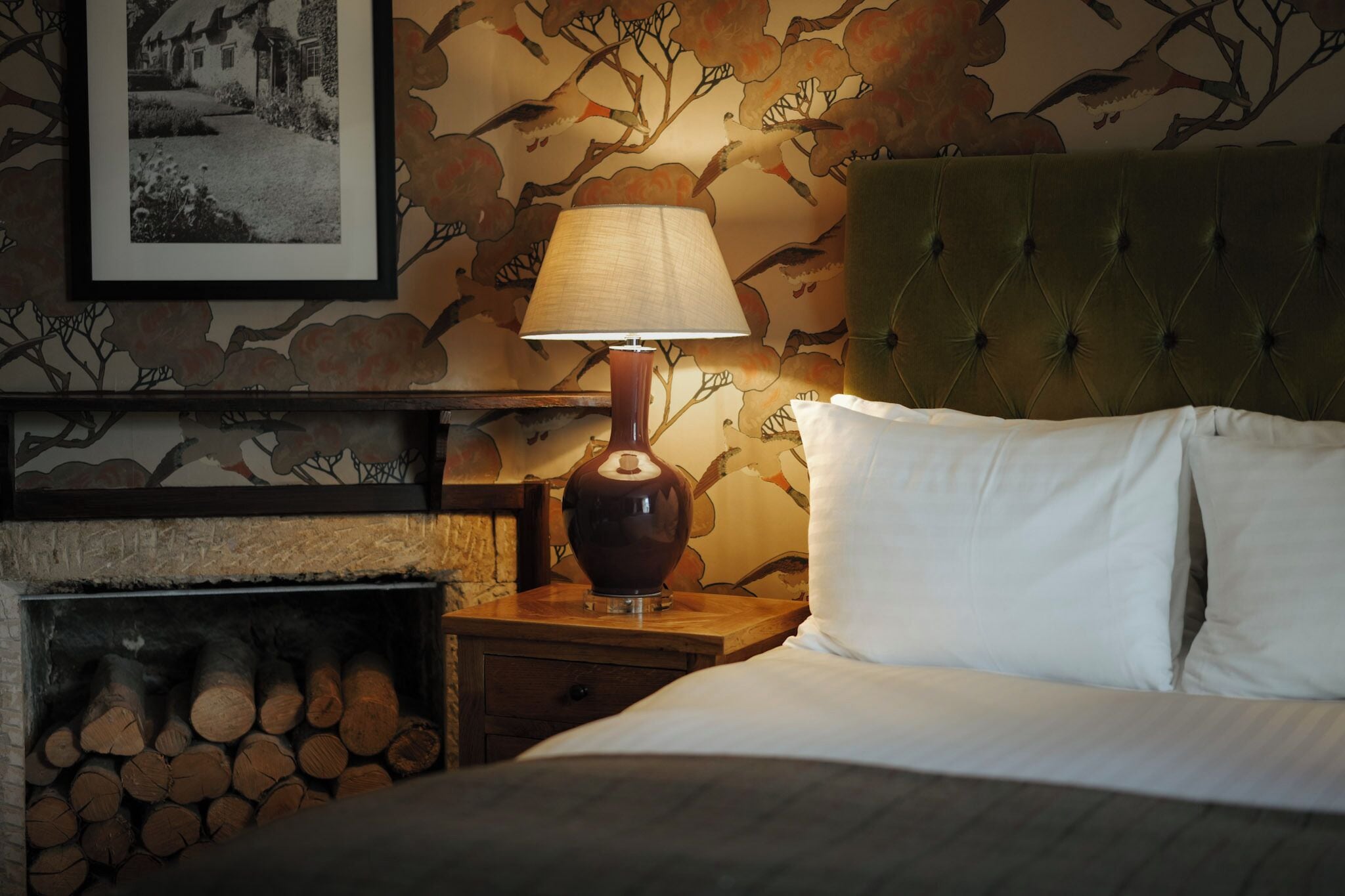 Une chambre à coucher avec un lit, une table de chevet avec une lampe, une cheminée avec des bûches et un mur orné d’un motif floral.