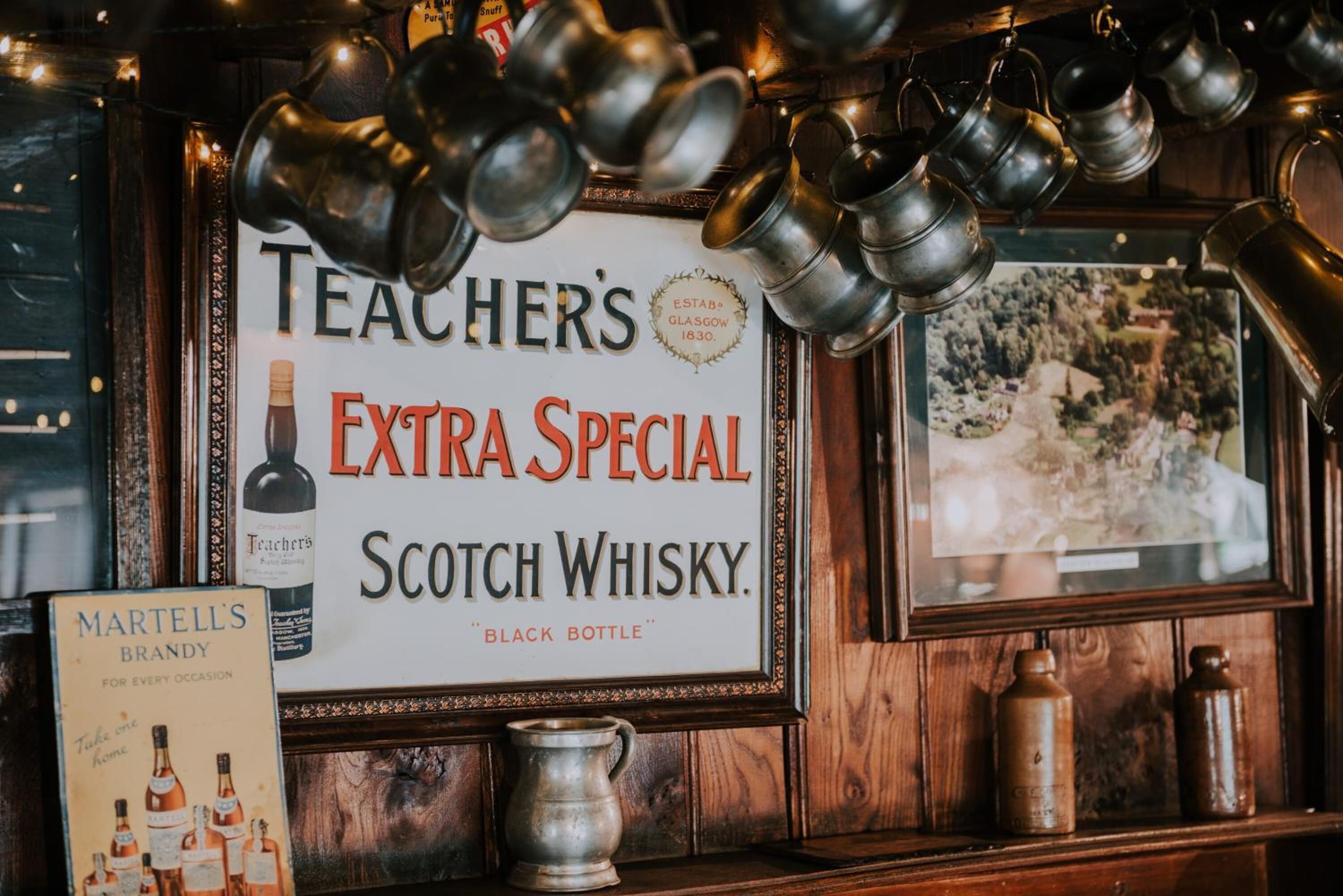Un letrero enmarco para el whisky escocés Teacher's Extra Special, un libro titulado 'Martell's Brandy' y varias tazas metálicas sobre una estantería de madera.