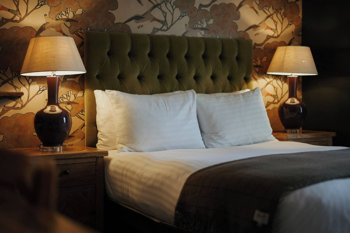 A neatly made bed with a tufted headboard, two bedside lamps, and a floral wallpaper pattern.
