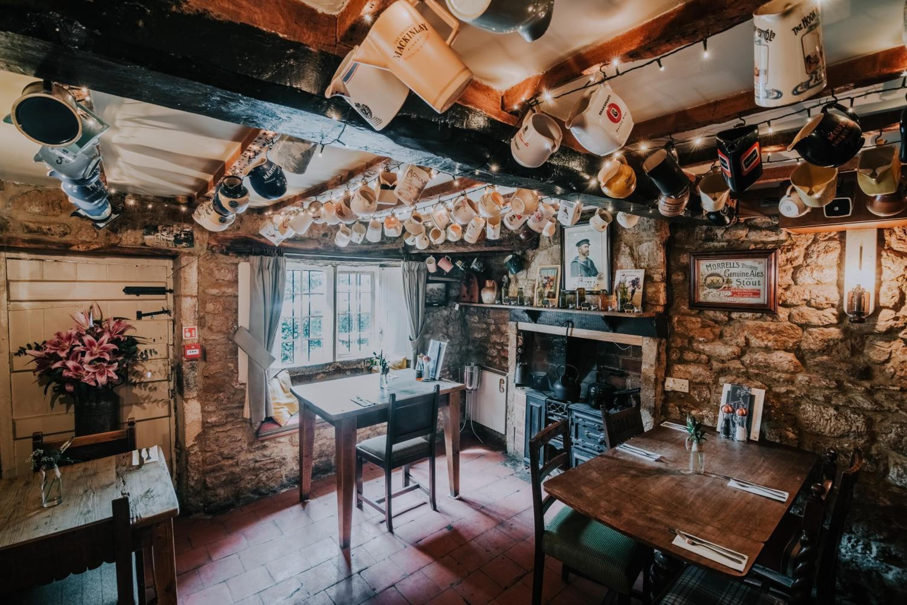 Un pub rústico con paredes de piedra vista, vigas de madera y una variedad de jarras colgando del techo.