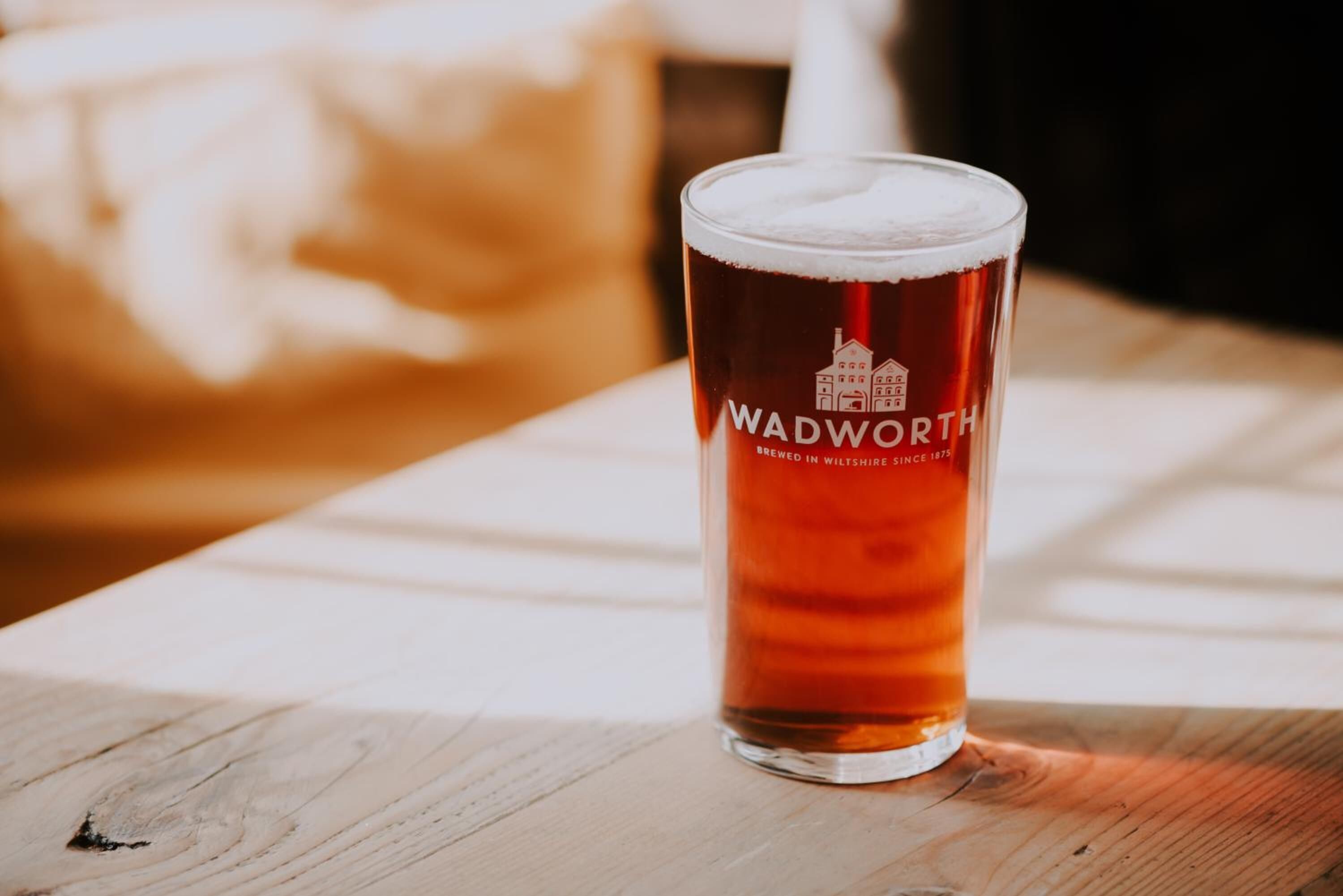 A glass of beer with the label "WADWORTH BREWED IN WILTSHIRE SINCE 1875" on a wooden table.