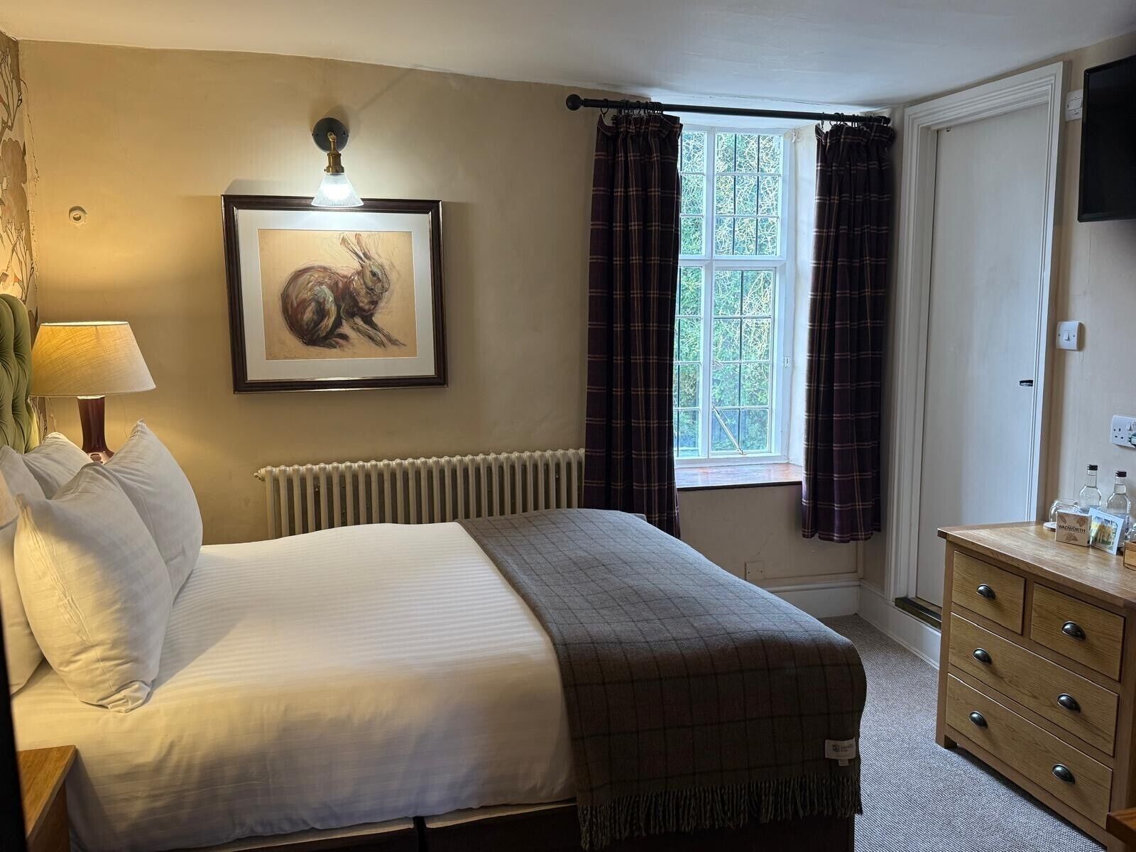 A bedroom with a bed, a radiator, a framed picture, a window with curtains, a dresser, and a lamp.