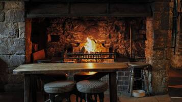 Una chimenea de piedra con fuego, una mesa de madera y dos taburetes en un interior rústico.