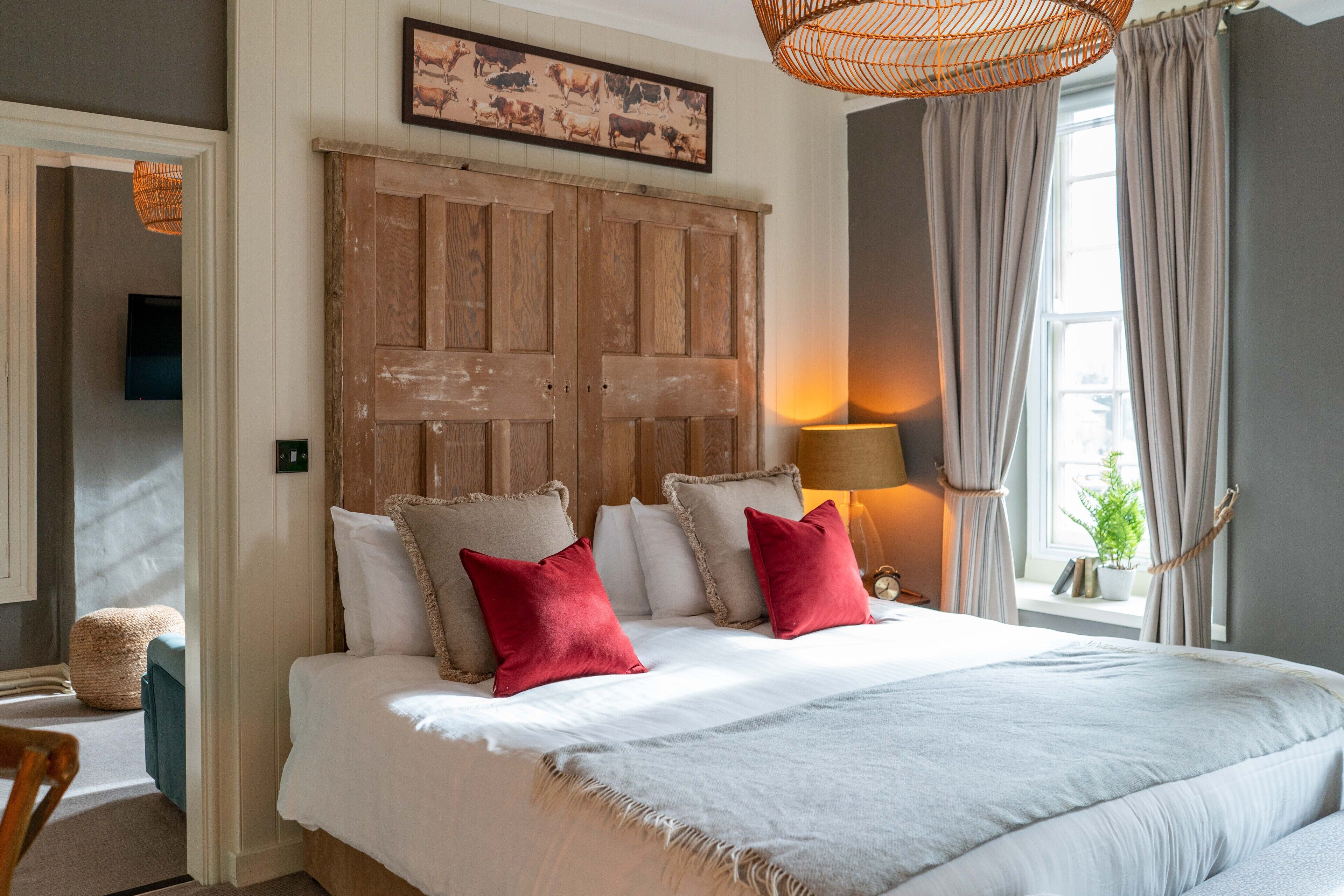 A bedroom with a large bed, two red pillows, a wooden headboard, a wall-mounted TV, and a window with curtains.