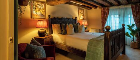 A bedroom with a wooden bed, a nightstand with a lamp, a chair, and a window with curtains.