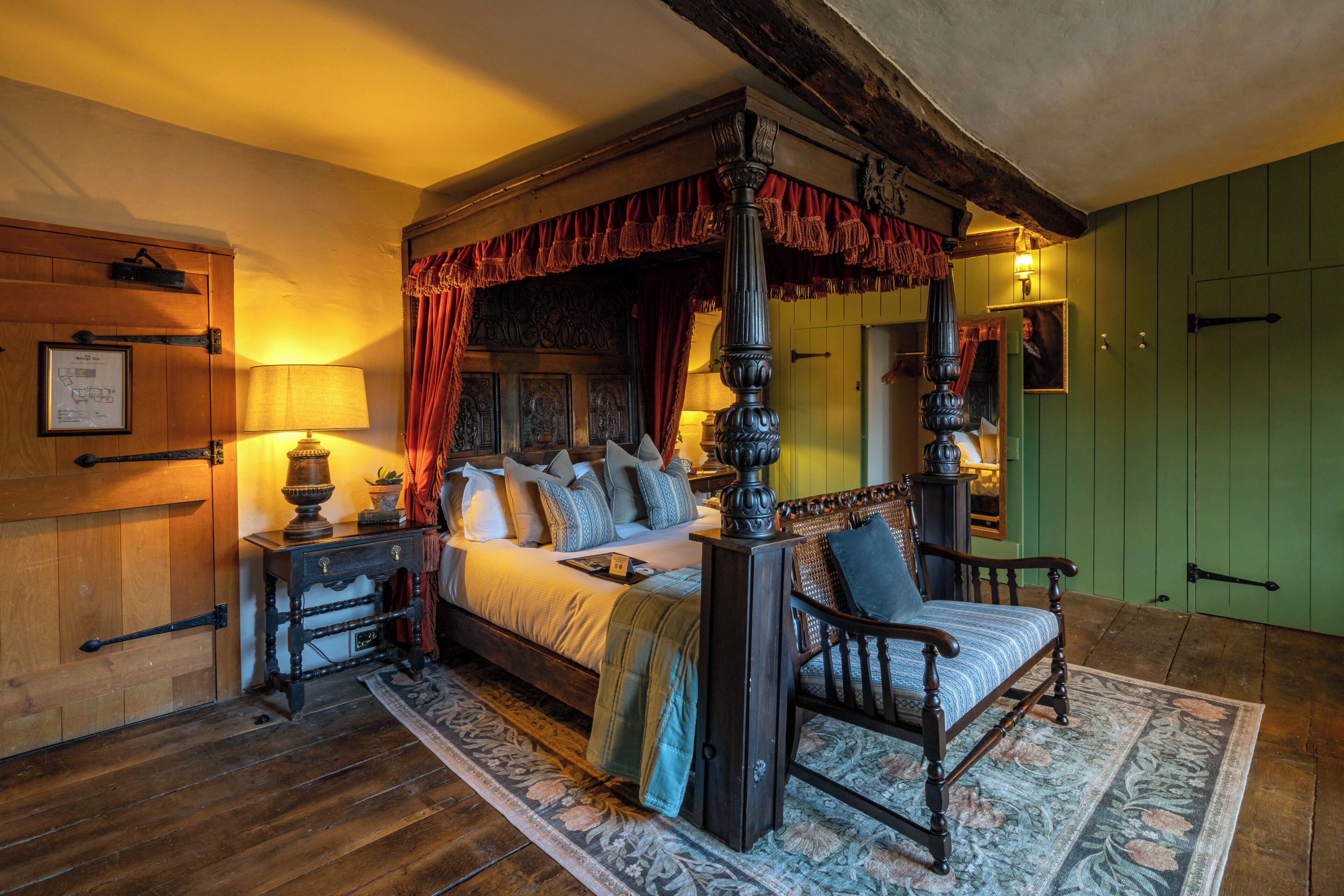 Una cama con dosel y cuatro columnas, una mesita de noche de madera, una silla y una puerta verde en una habitación con suelo de madera.