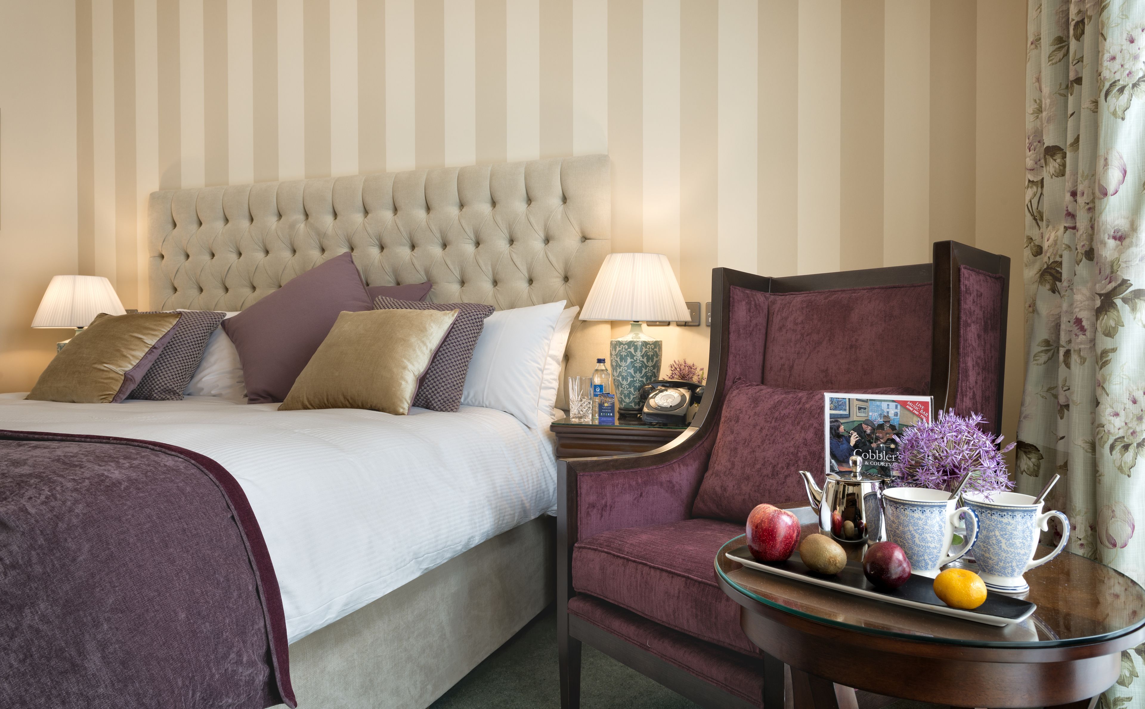 A hotel room with a large bed, a nightstand, a table with fruits and a teapot, and a floral curtain.