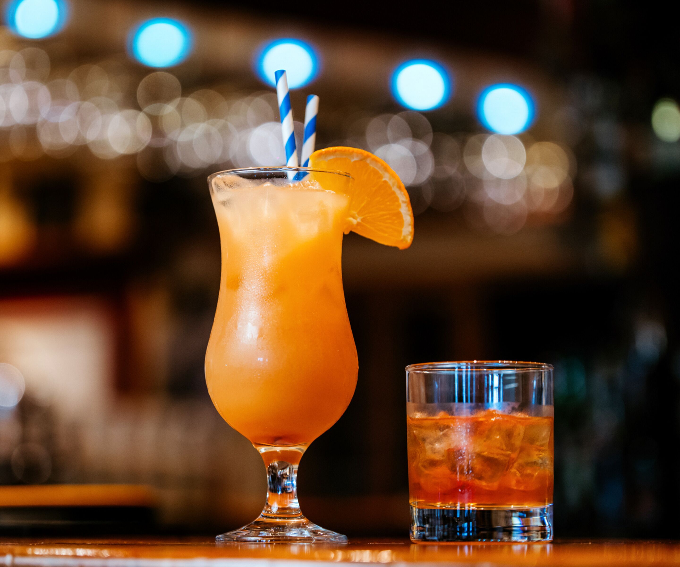 Un bar avec un grand verre contenant une boisson orange, décorée d’une tranche d’orange et de pailles rayées, et un verre plus petit contenant une boisson similaire.