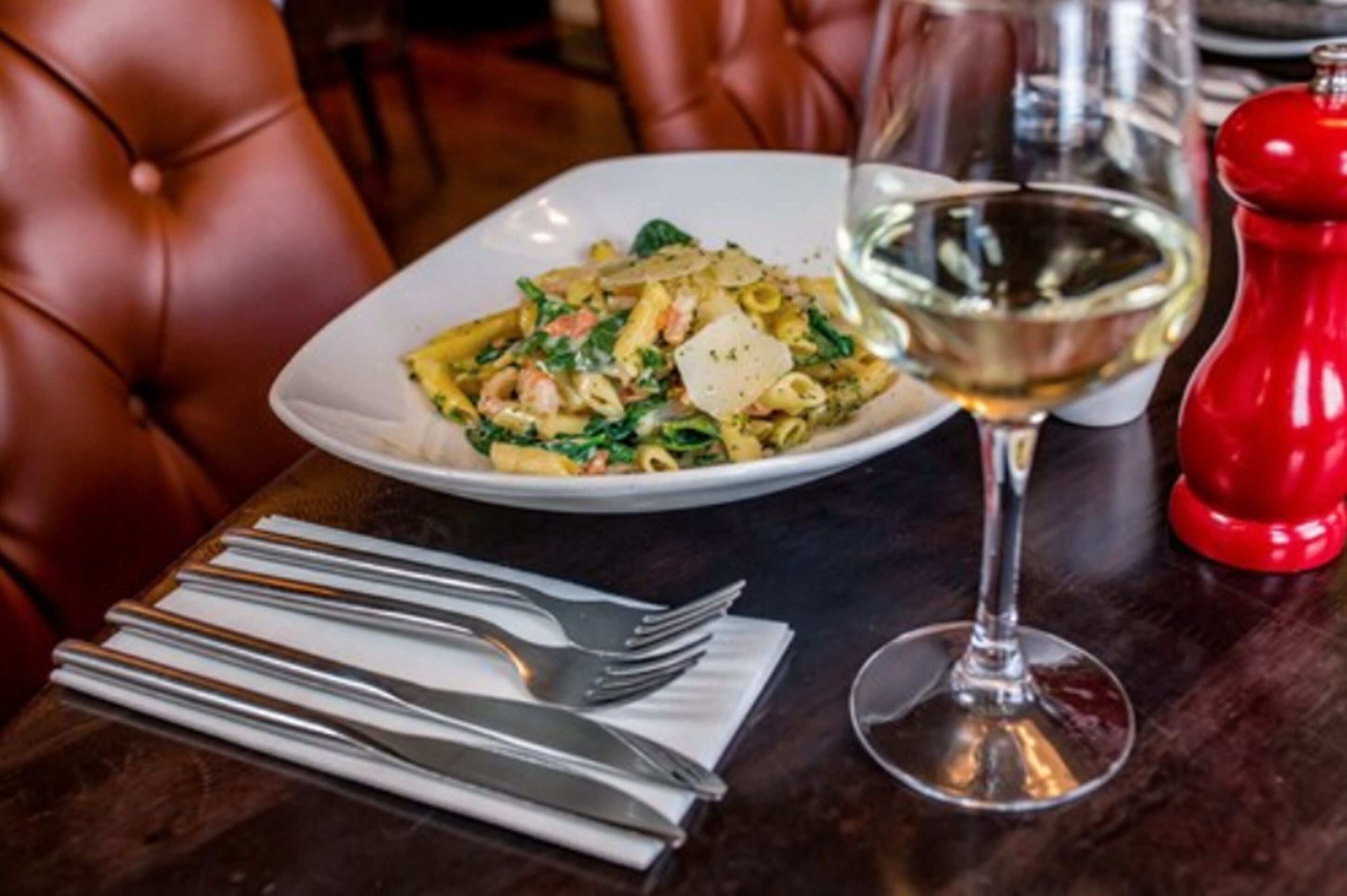 A dining table with a plate of pasta, a glass of white wine, and a red pepper grinder.