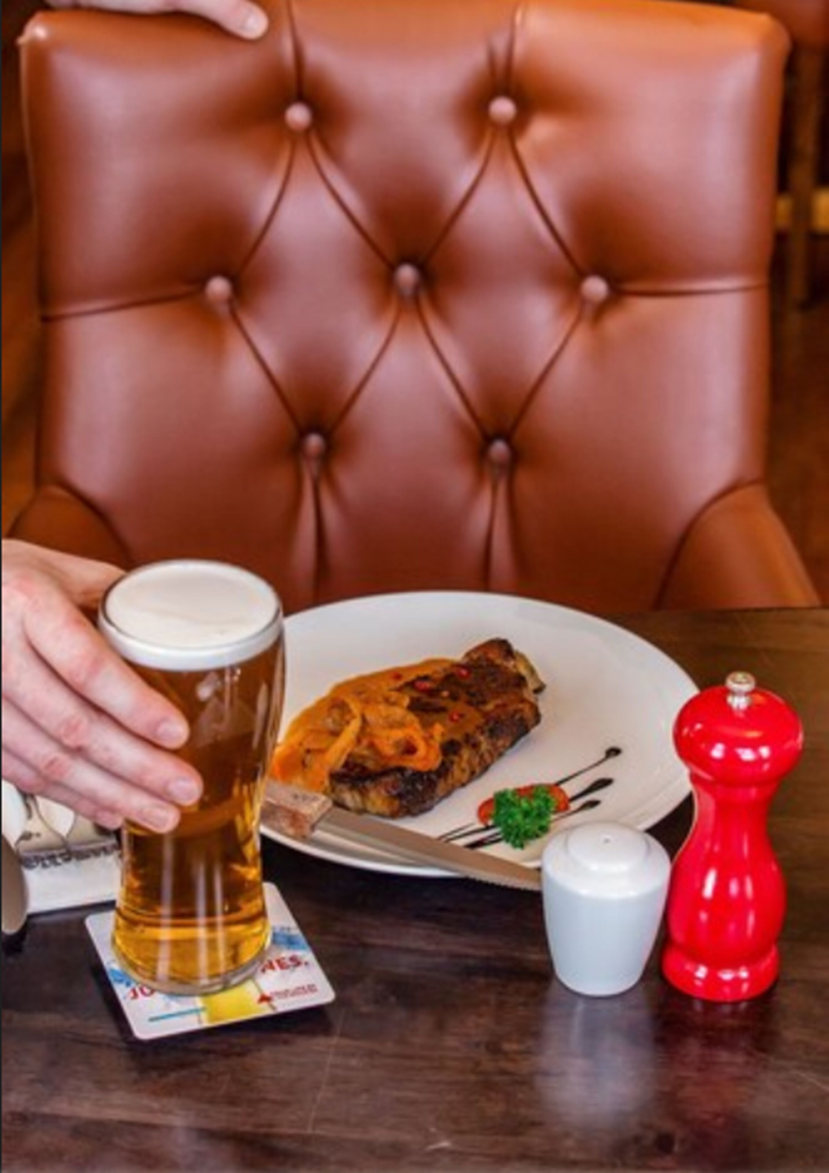 A dining table with a plate of food, a glass of beer, and a red pepper grinder.