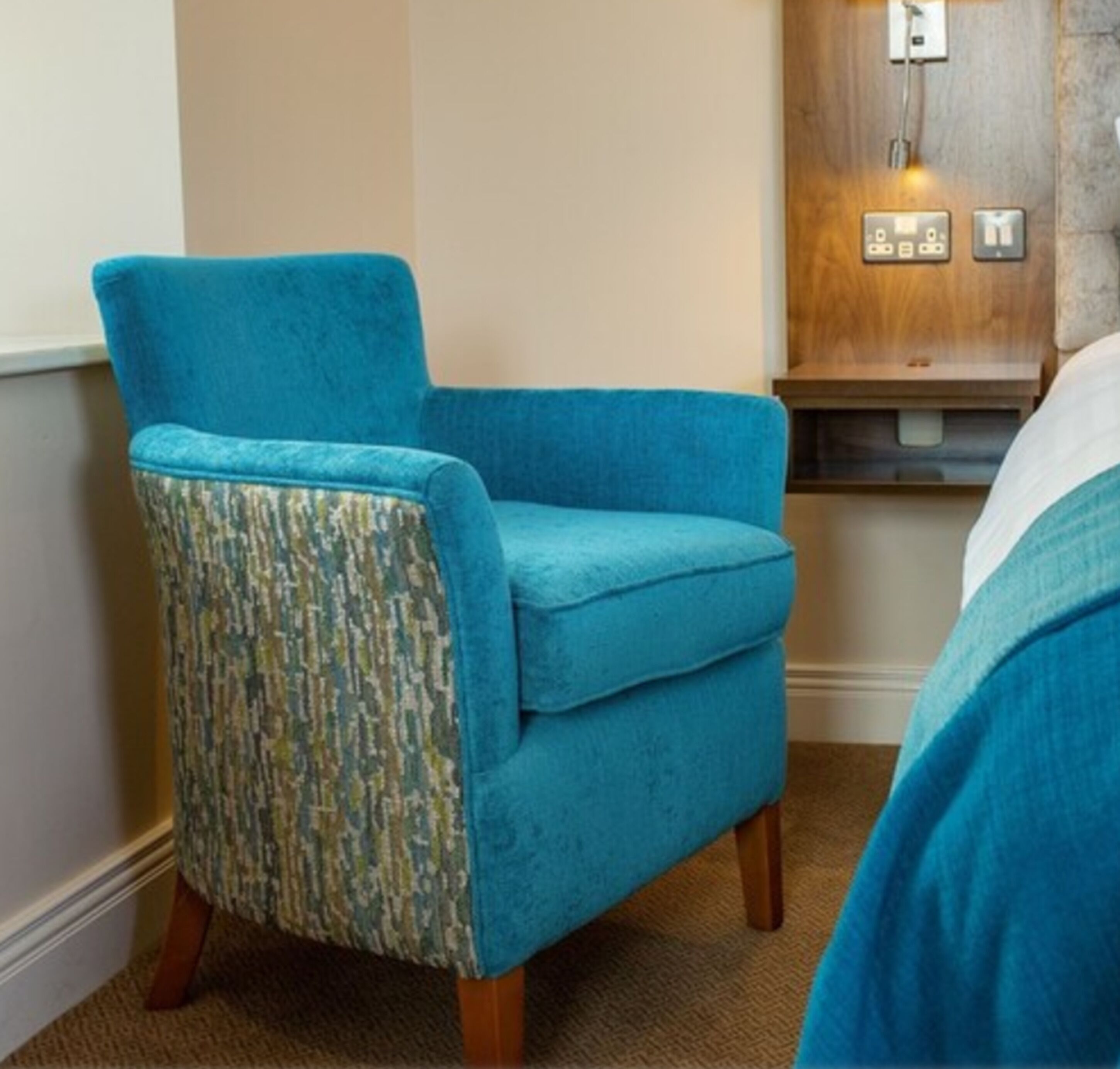 A blue armchair with a patterned backrest in a room with a wooden wall panel and a bed with white bedding.