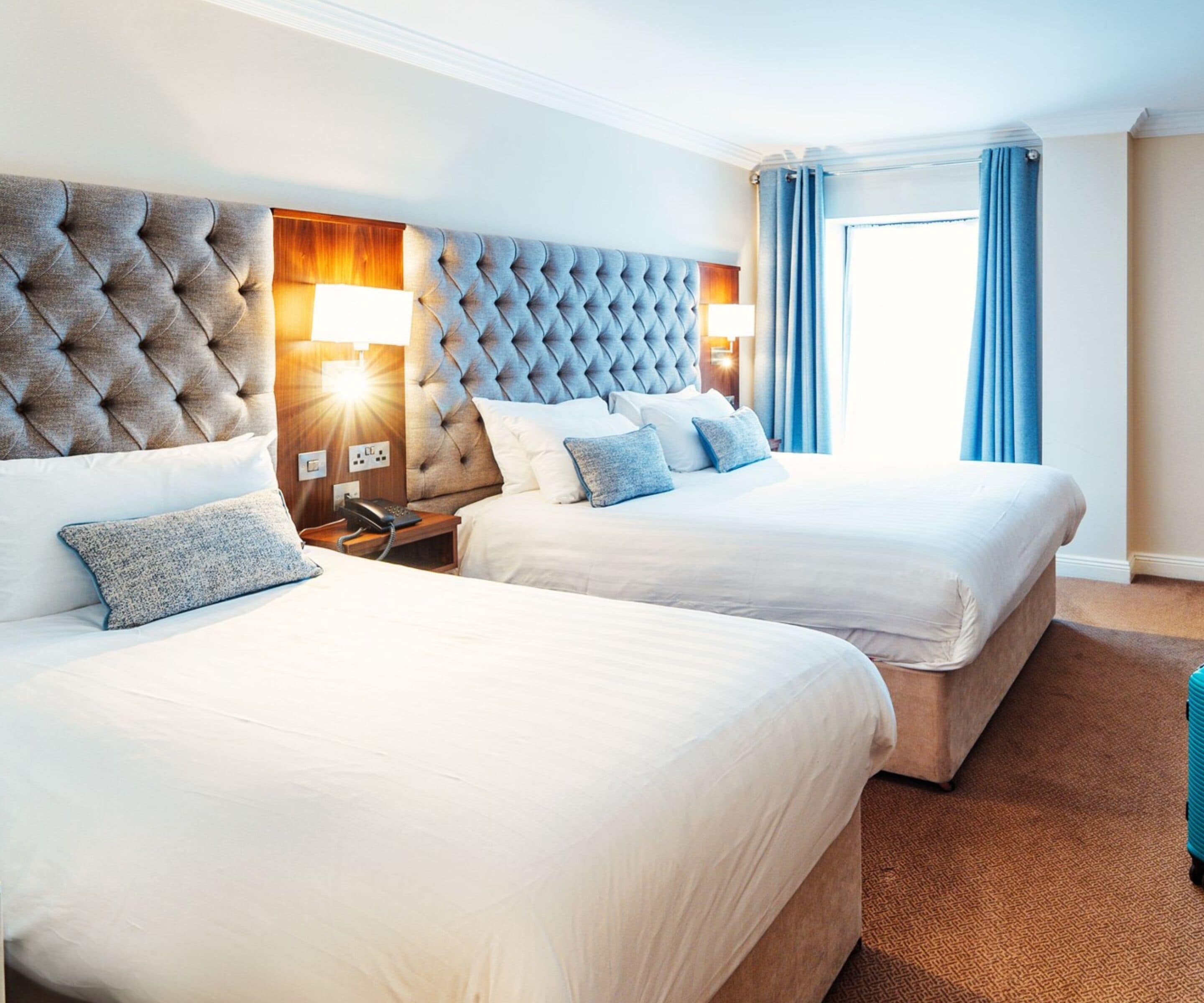 A hotel room with two beds, a tufted headboard, bedside lamps, and a view of the outdoors.