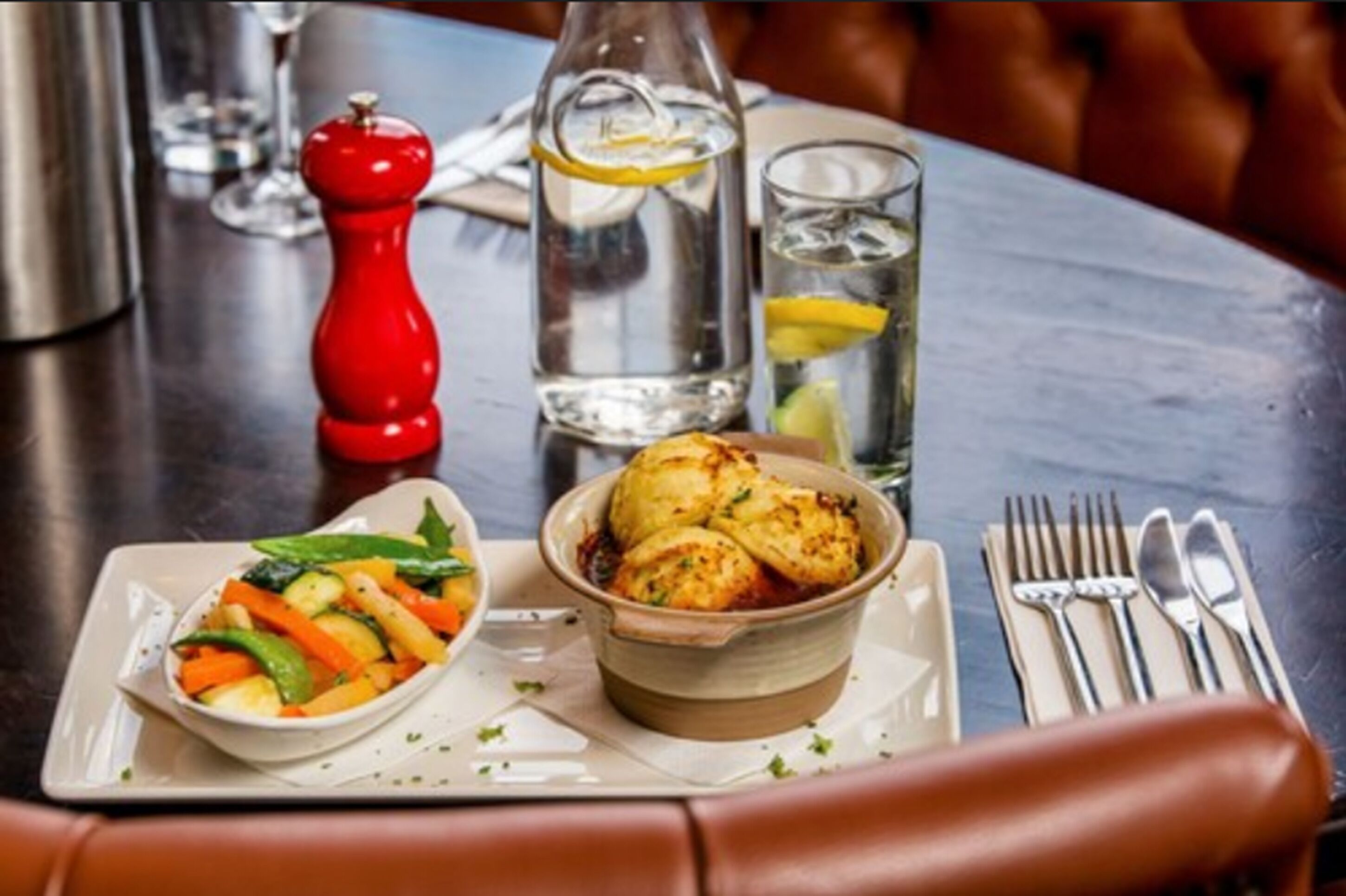 A dining table with a plate of food, a bottle of water, a glass of water, a red pepper shaker, and cutlery.