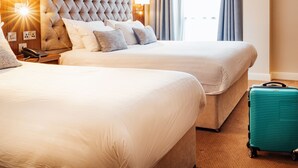 Quarto de hotel com duas camas, uma cabeceira grande com efeito acolchoado, luminárias laterais, um telefone e uma mala.