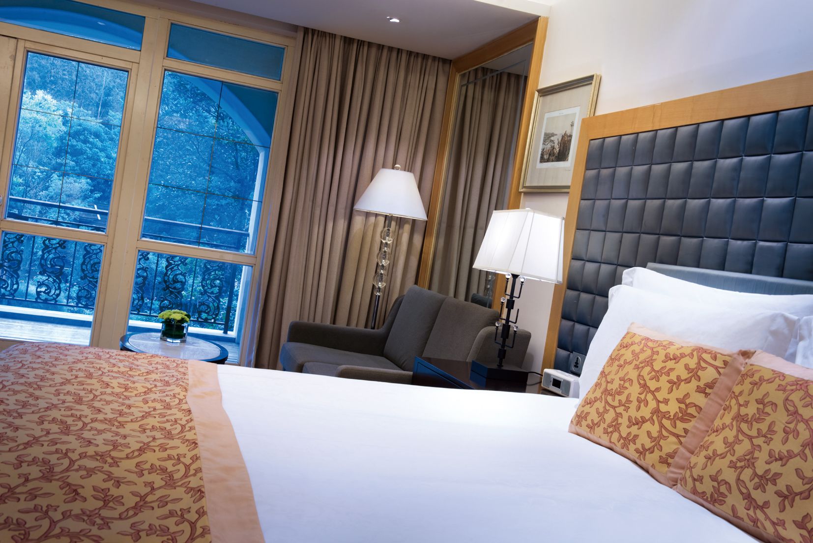 A hotel room with a large bed, a chair, a small table with a lamp, and a window with curtains.