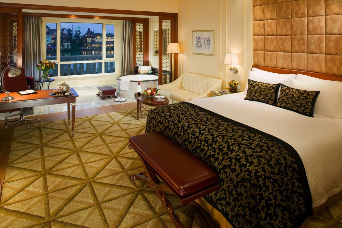 Een hotelkamer met een groot bed, een bureau met een stoel, een kleine tafel met een bloemenvaas, een bankje en een stadsgezicht door grote ramen.
