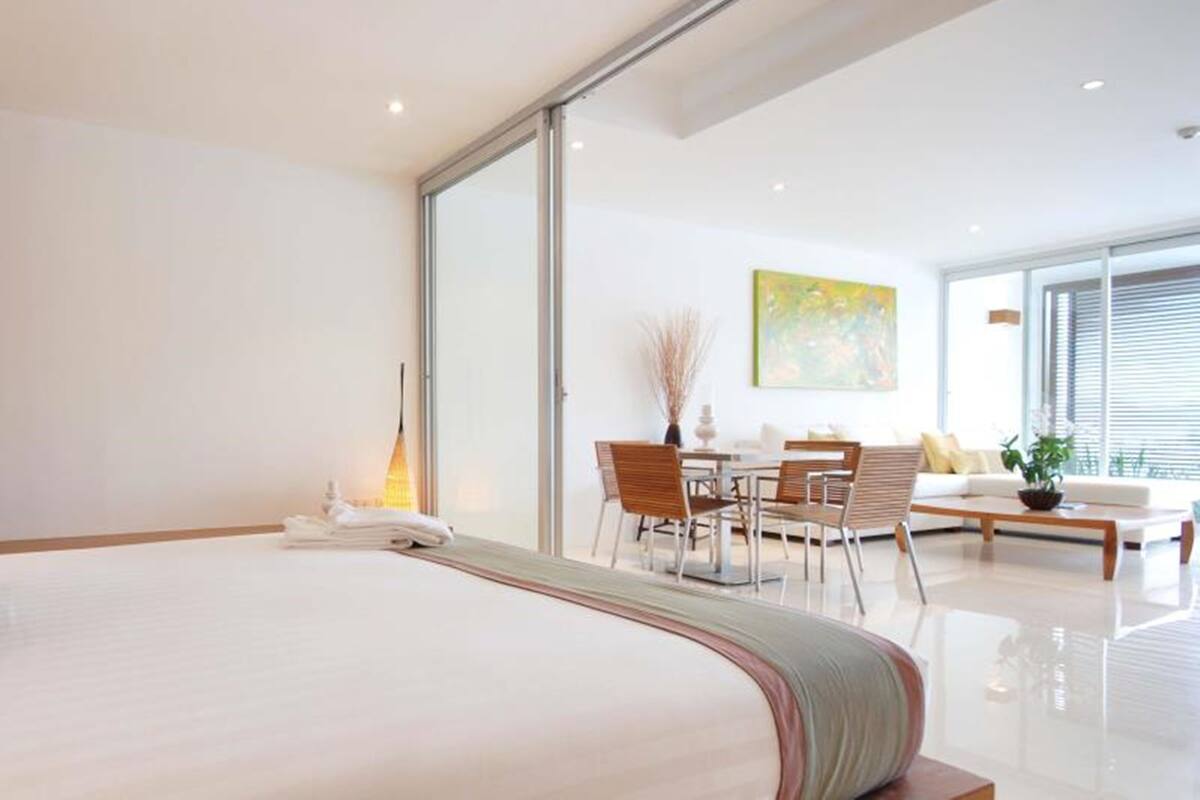 Una habitación de hotel moderna con una cama grande, un área de comedor con una mesa y sillas, un sofá y un ventanal con persianas.