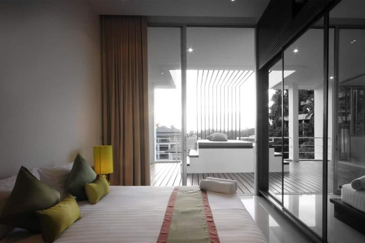 Una habitación de hotel moderna con una cama grande, una mesita de noche, una lámpara y vistas a un balcón con barandilla.