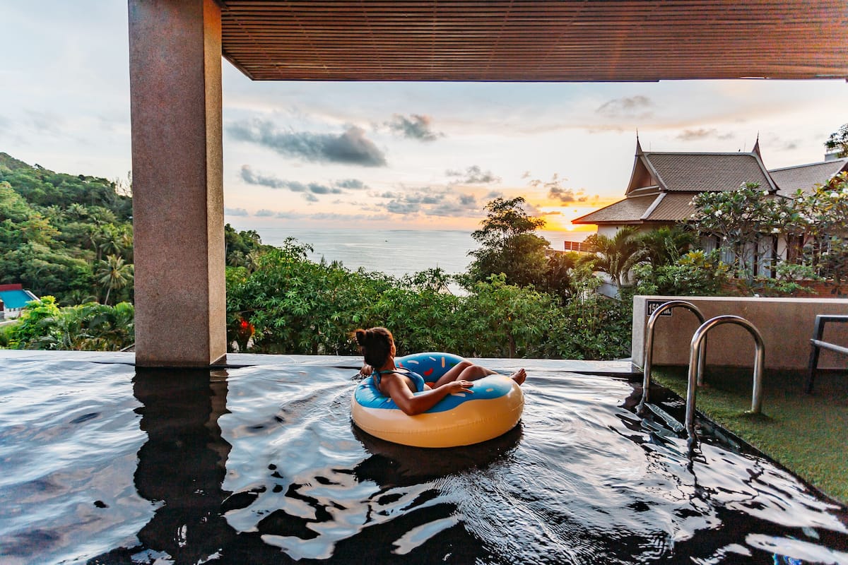 A person floating on an inflatable tube in a pool with a view of a traditional building and lush greenery in the background.