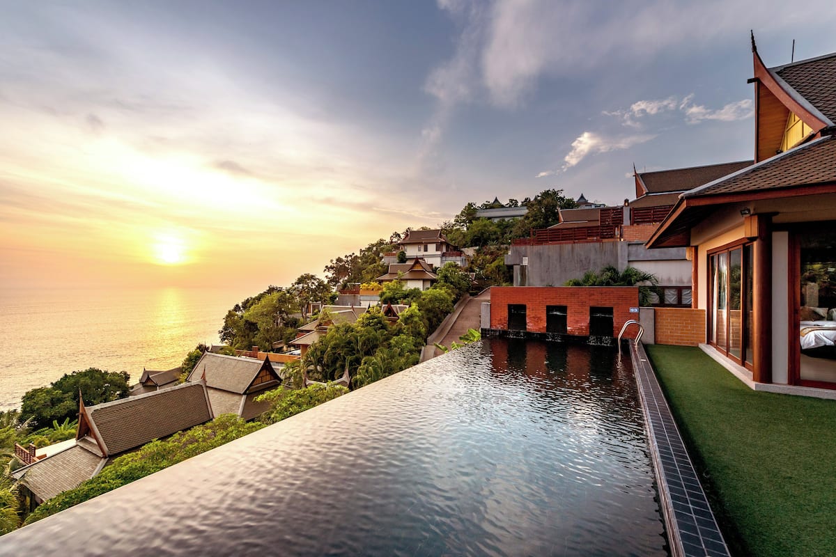 A swimming pool with a view of the sunset over the sea, surrounded by traditional houses with a modern building on the right.