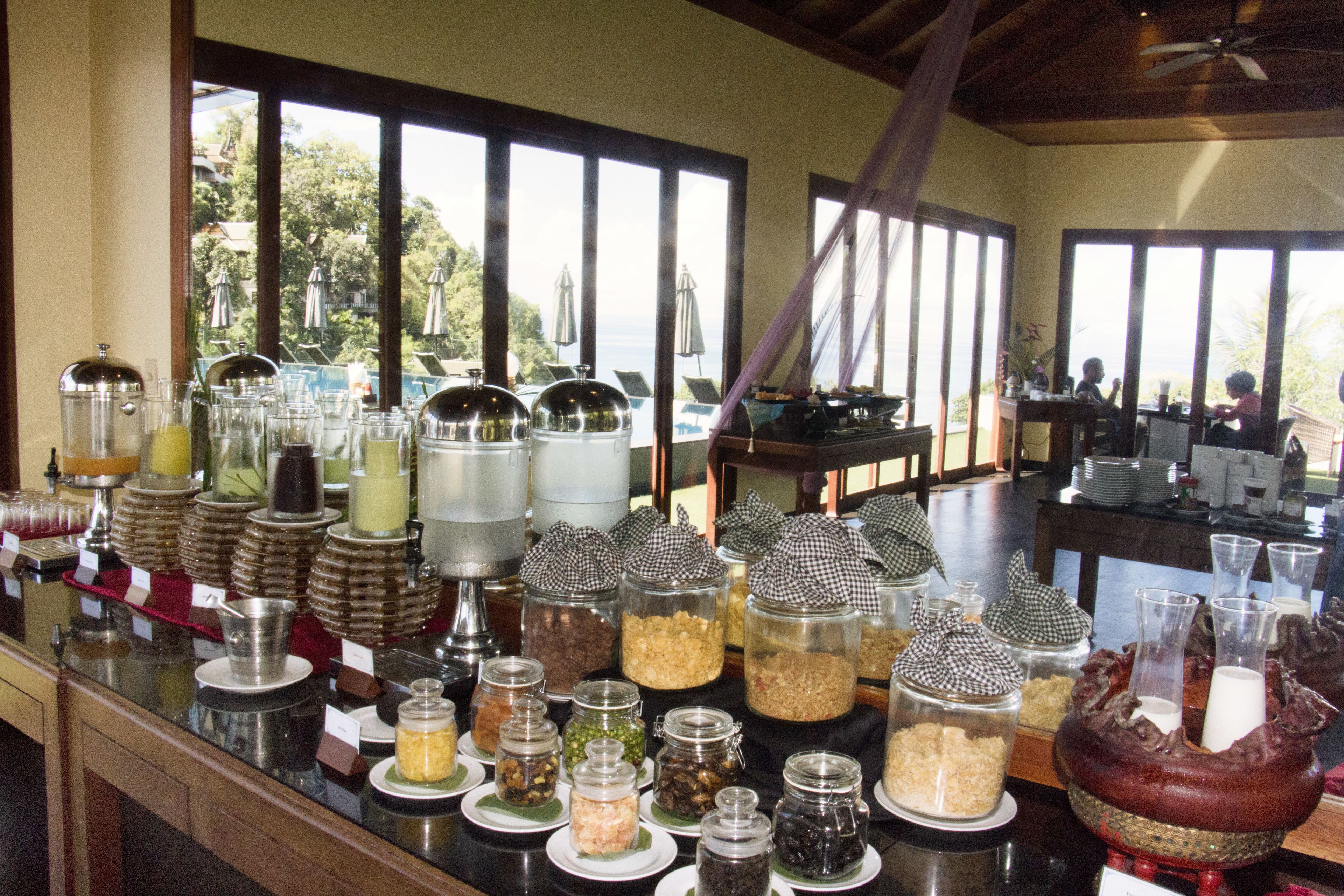 Un espace buffet proposant divers plats présentés dans des récipients en verre, un ventilateur de plafond et de grandes fenêtres offrant une vue sur l’extérieur.