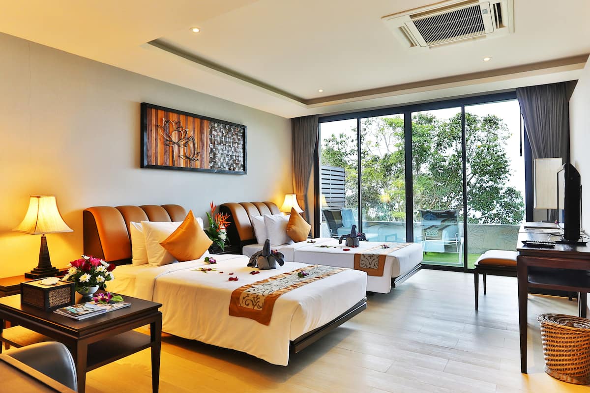 A hotel room with a large bed, a smaller bed, a desk, and a chair. There is a window with a view of trees and a balcony.