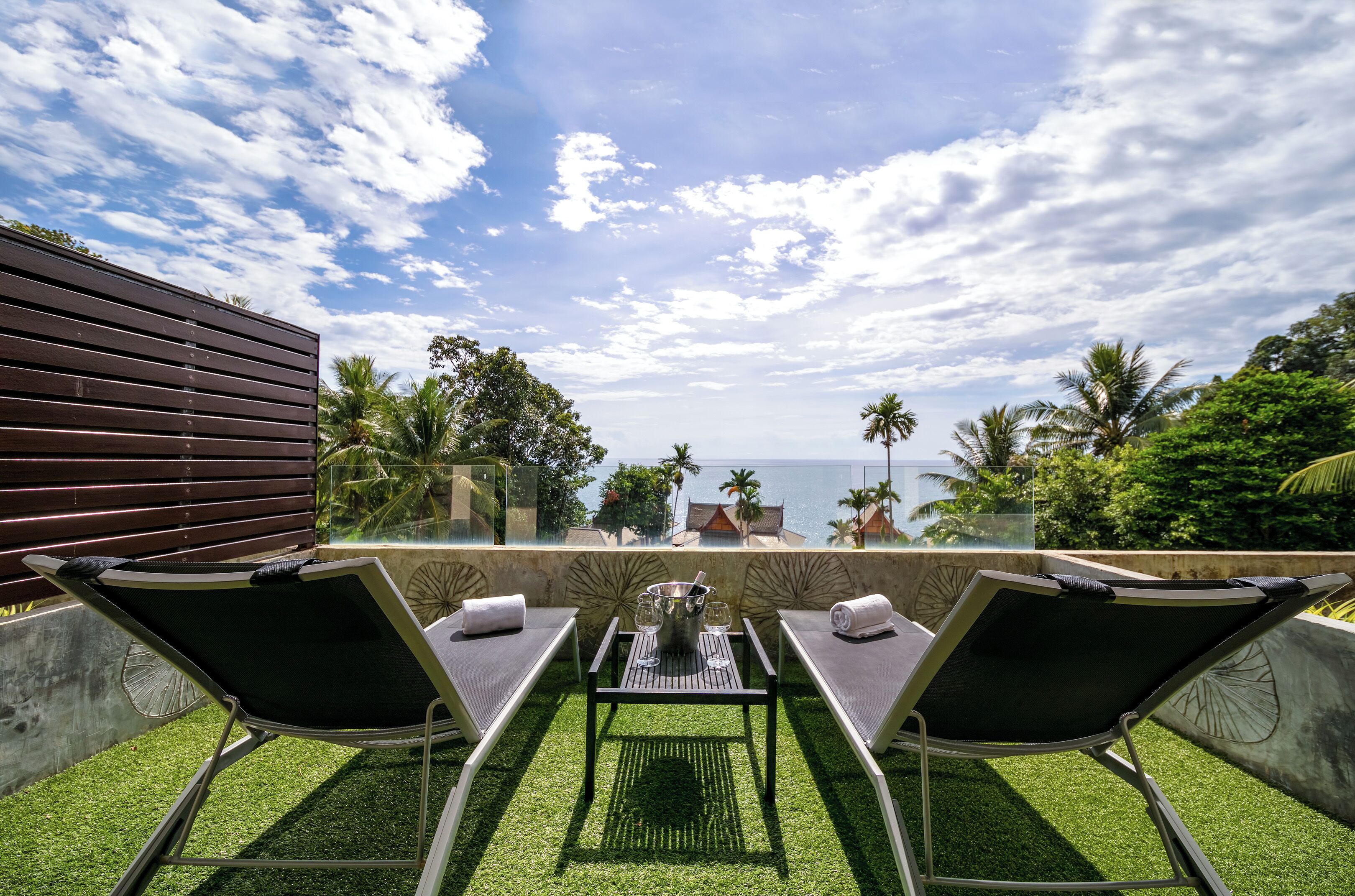 A terrace with two lounge chairs, a table with a drink tray, and a view of the sea and palm trees.