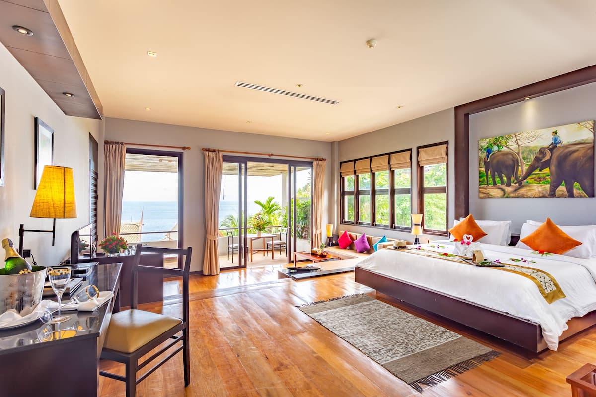 A hotel room with a large bed, a dining area, a balcony with a view, and a painting on the wall.