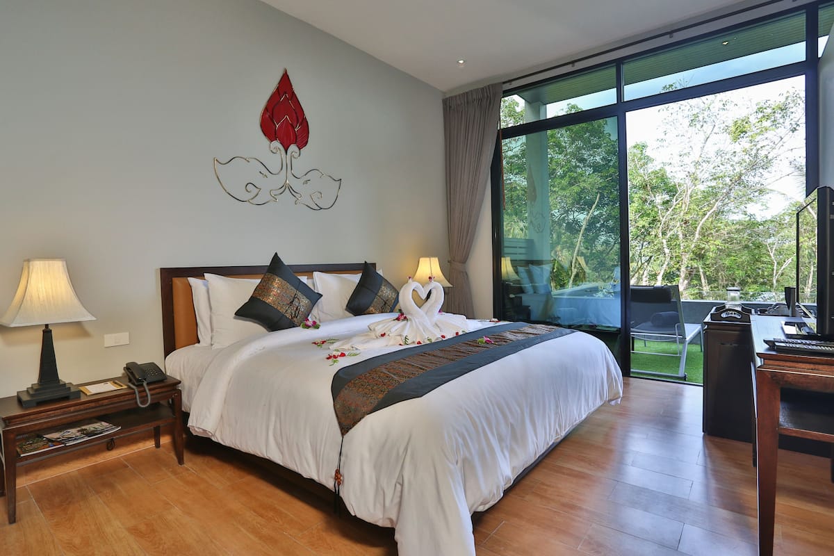 A hotel room with a large bed, two bedside lamps, a desk with a chair, and a view of the outdoors through large windows.