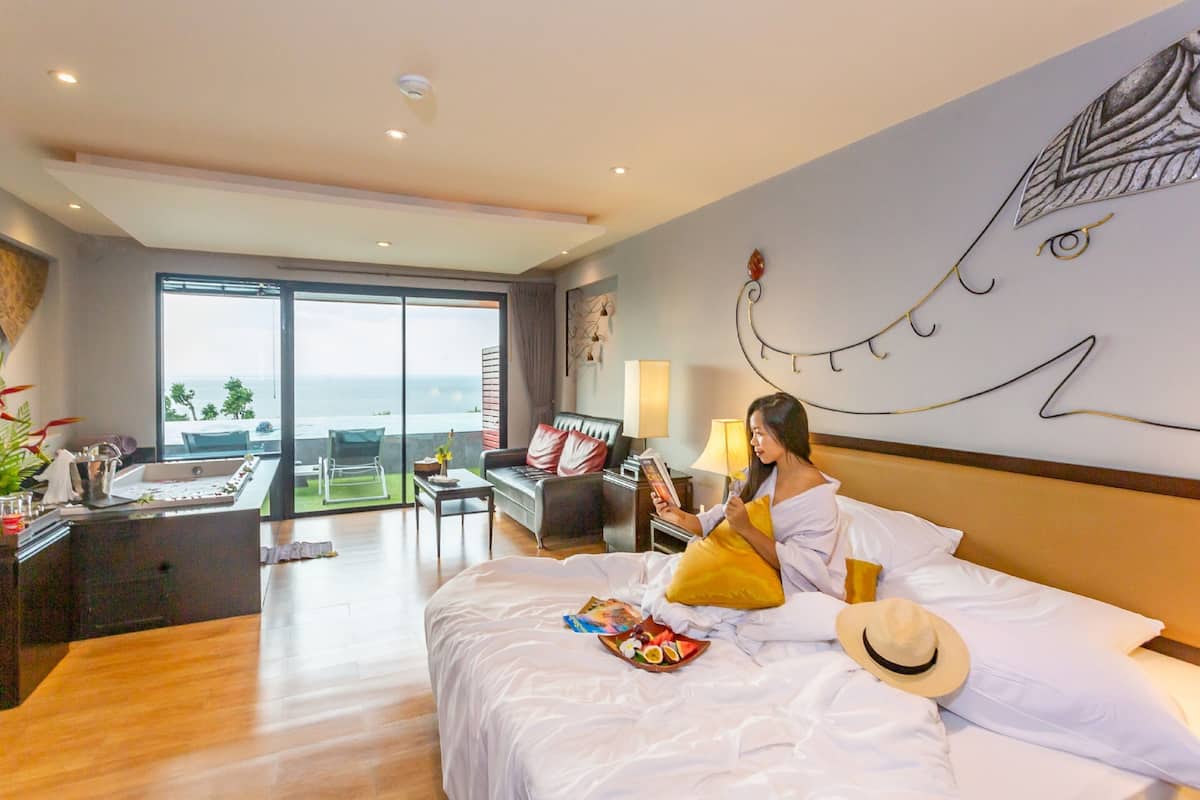 A hotel room with a bed, a reading area with a sofa and a table, a large window with a view of the sea, and a wall art piece.