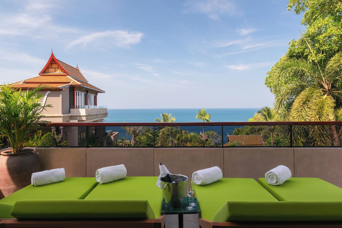 A balcony with two green cushioned chairs, a small table with a bucket and glasses, and a view of the ocean and a traditional building.