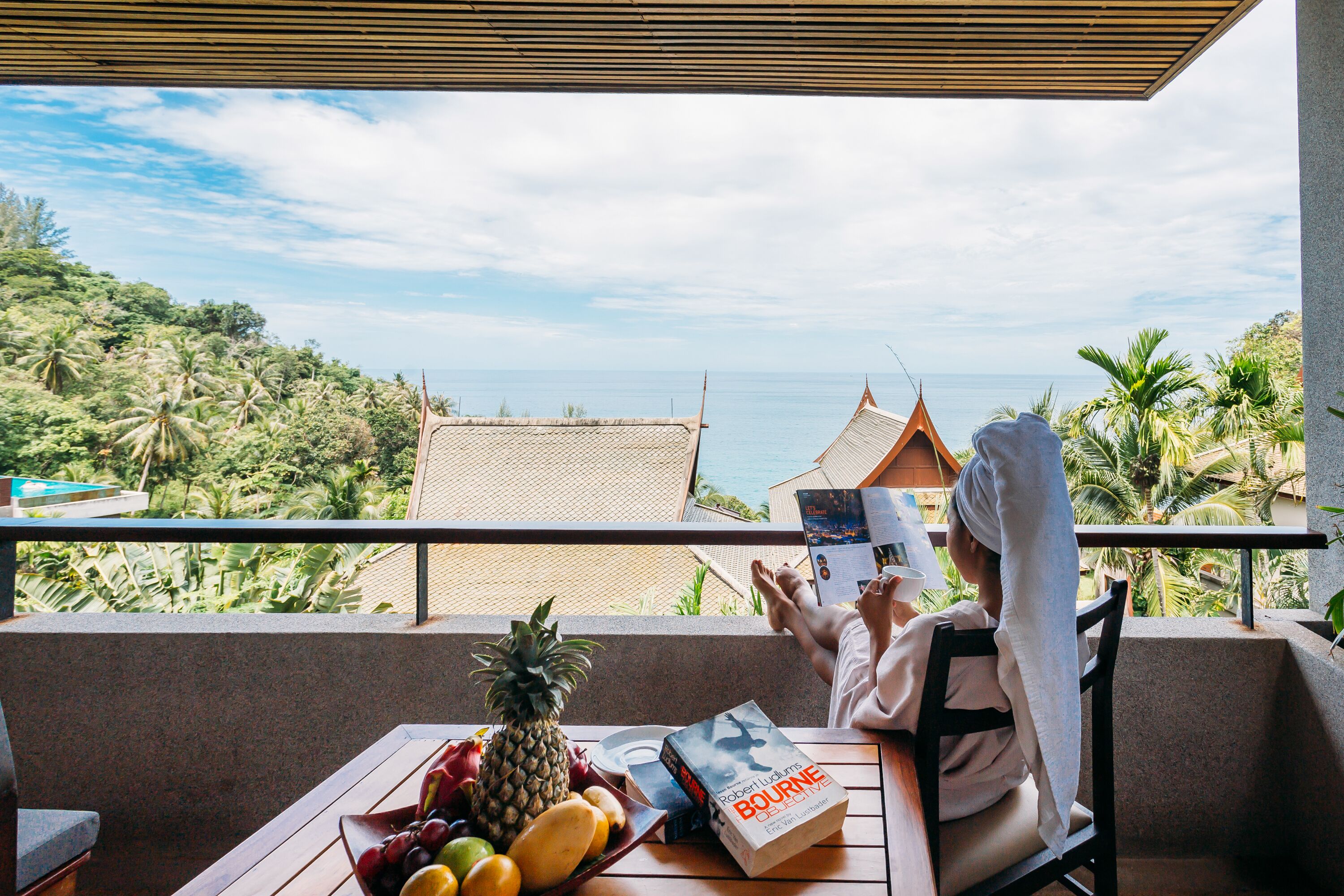 A person reading a book on a balcony with a view of the sea and palm trees.