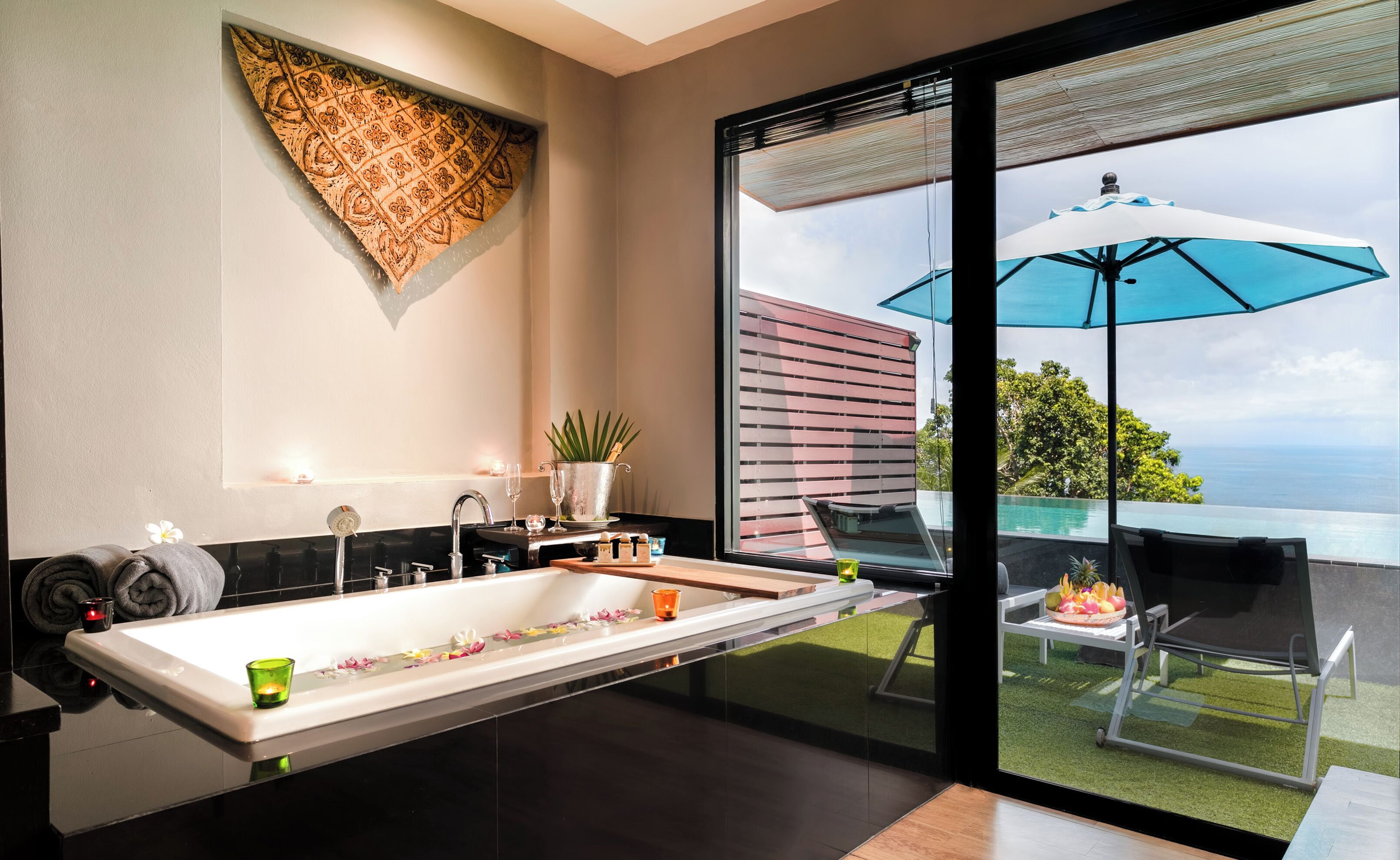 Un baño moderno con una bañera grande, un lavamanos y una cabina de ducha con mampara de vidrio. Hay un ventanal con vista al océano y un área de patio con mobiliario exterior.