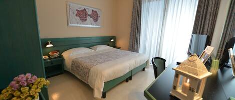 A hotel room with a bed, a nightstand, a desk, a chair, a vase of flowers, a painting, and a window with curtains.