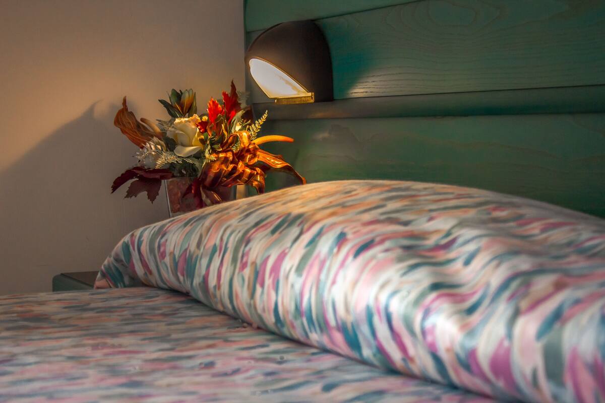 A bed with a colorful patterned bedspread, a bedside table with a vase of flowers, and a wall-mounted light fixture.