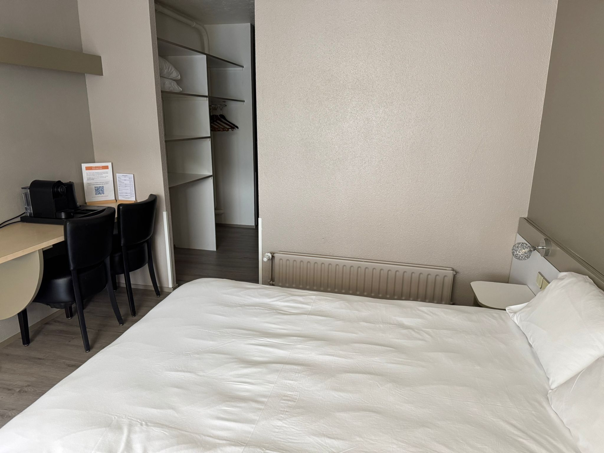 Habitación de hotel con cama, escritorio, silla, radiador y armario.