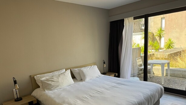 A hotel room with a large bed, a bedside table with a lamp, and a view of an outdoor patio with plants and a table. - Hotel Dupleix (Quimper)