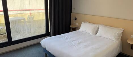 Habitación de hotel con una cama grande, una ventana con cortinas, una mesita de noche pequeña y vistas al exterior.