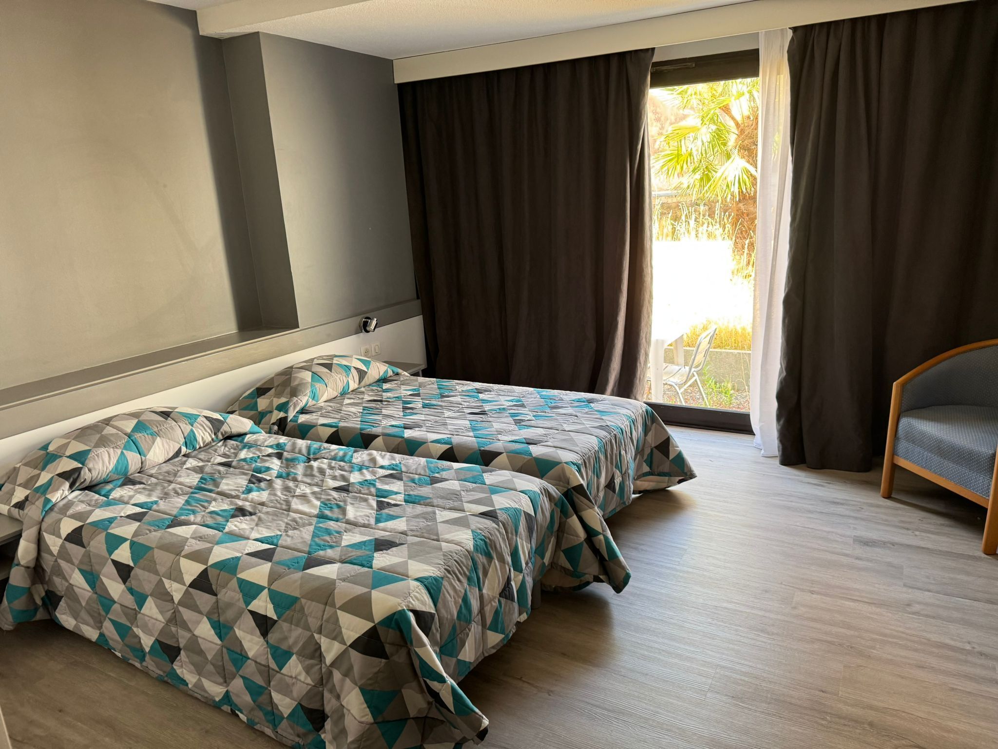 A hotel room with two beds, a wooden floor, and a view of palm trees through the window.