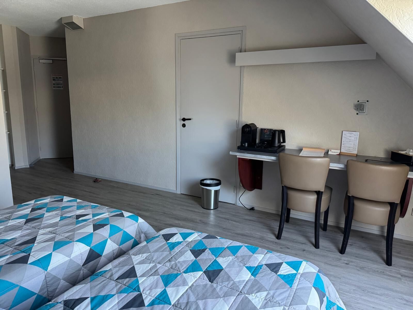 Quarto de hotel com cama, escrivaninha com cafeteira, duas cadeiras e um cesto de lixo.