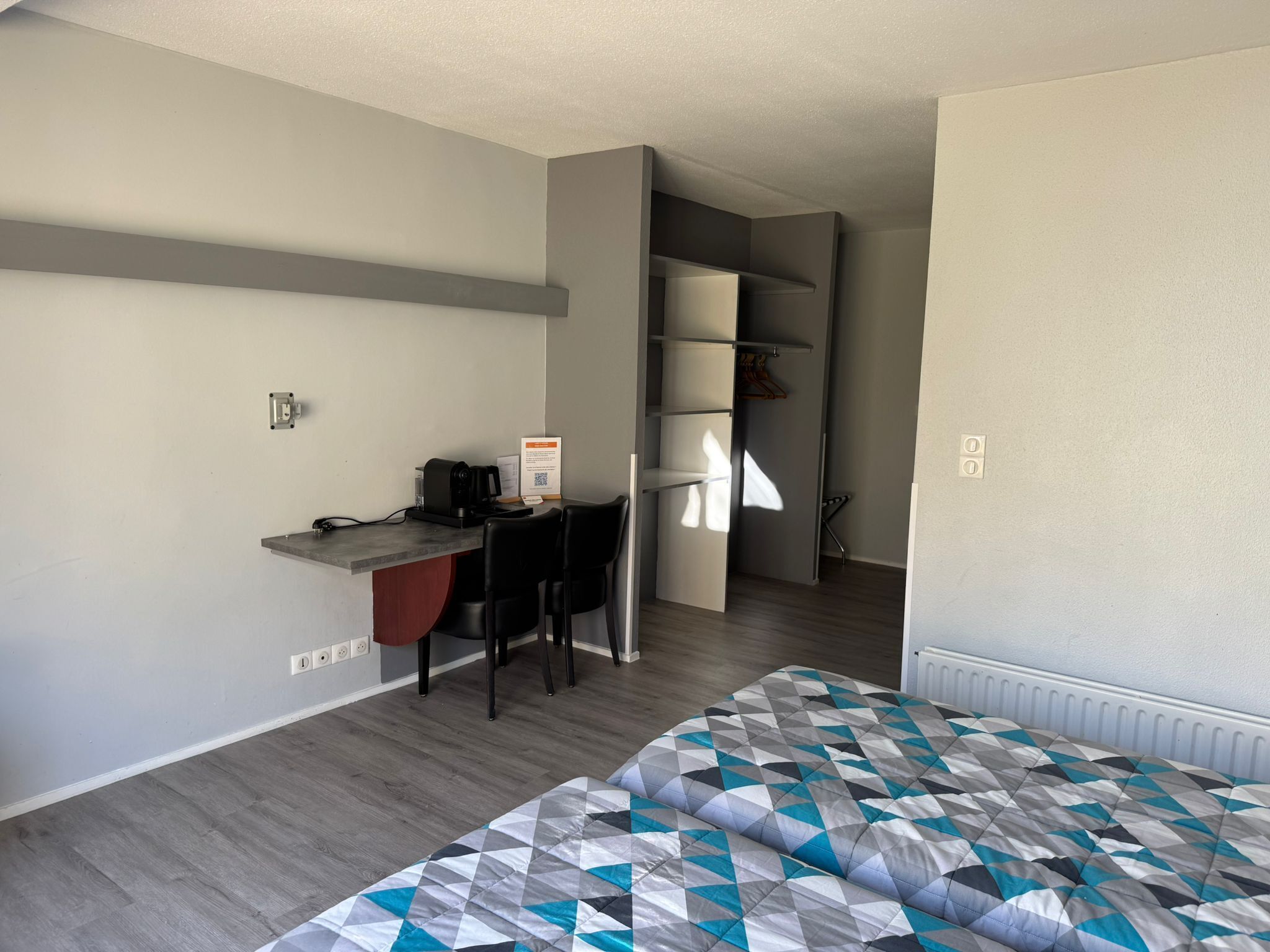 Une chambre d’hôtel de taille réduite comprenant un lit, un bureau avec une chaise, une petite kitchenette et une armoire.