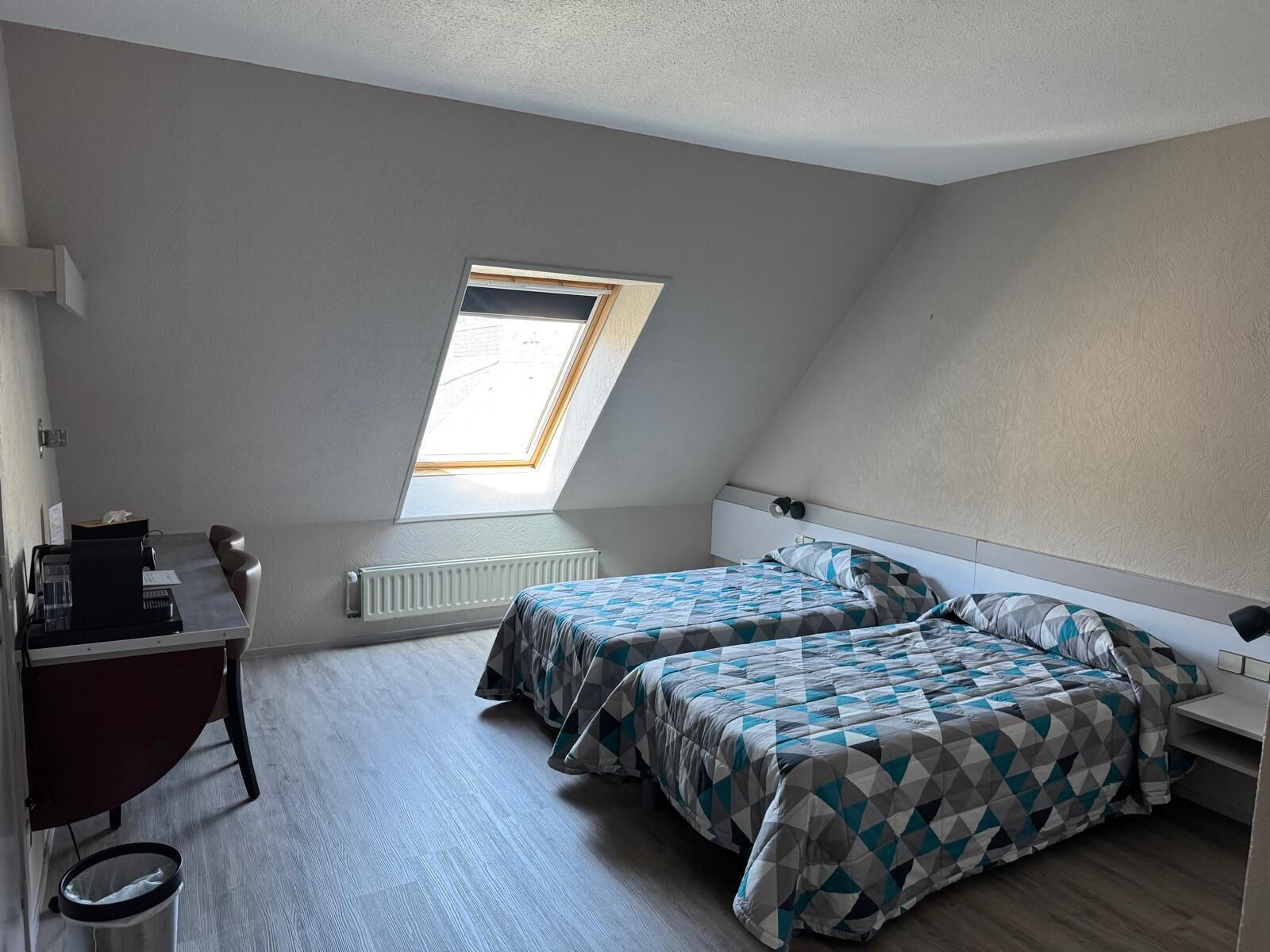 Une petite pièce bien éclairée, avec un plafond en pente, deux lits dotés de draps à motifs géométriques, un bureau avec un ordinateur et une fenêtre laissant entrer la lumière naturelle.