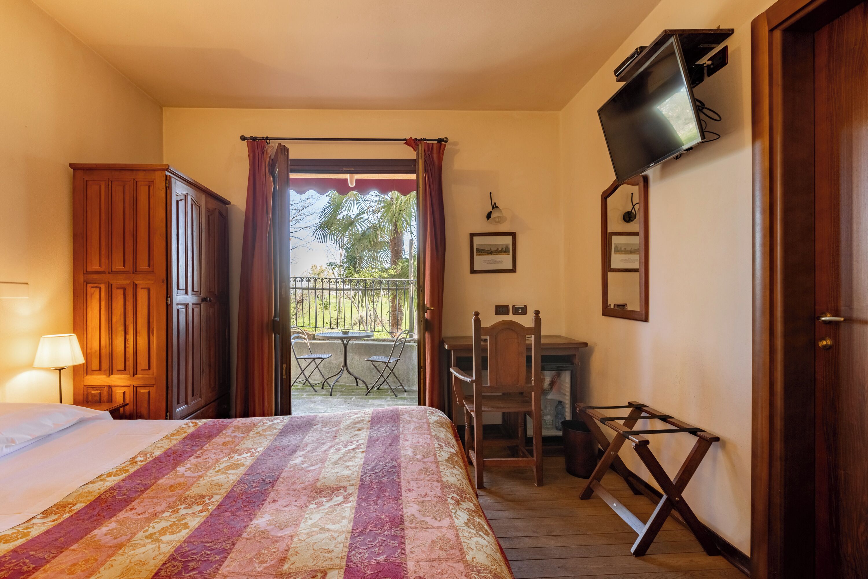 Een hotelkamer met een bed, een bureau, een televisie en een balkon met uitzicht op palmbomen.