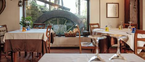 Un restaurante con mesas dispuestas para comer, un gran ventanal arqueado y una pared de piedra visible a través del ventanal.